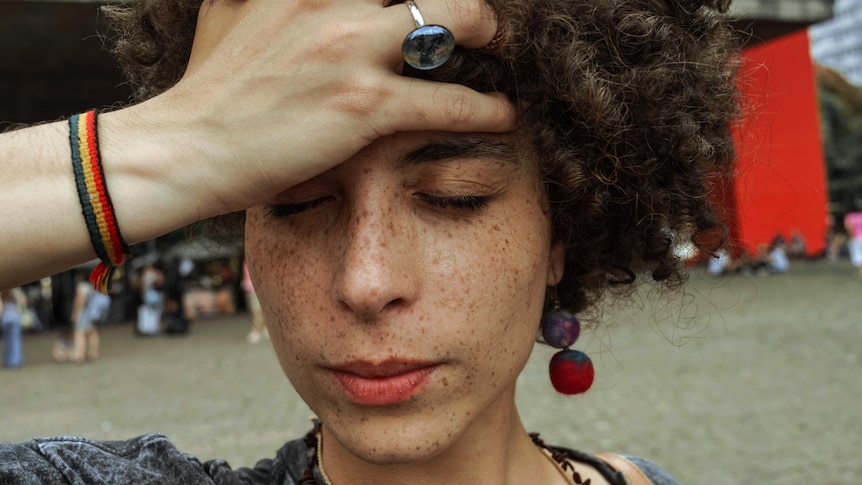 Close-up of young woman with curly brown hair and freckles, holding her hand to her forehead and frowning with eyes closed.