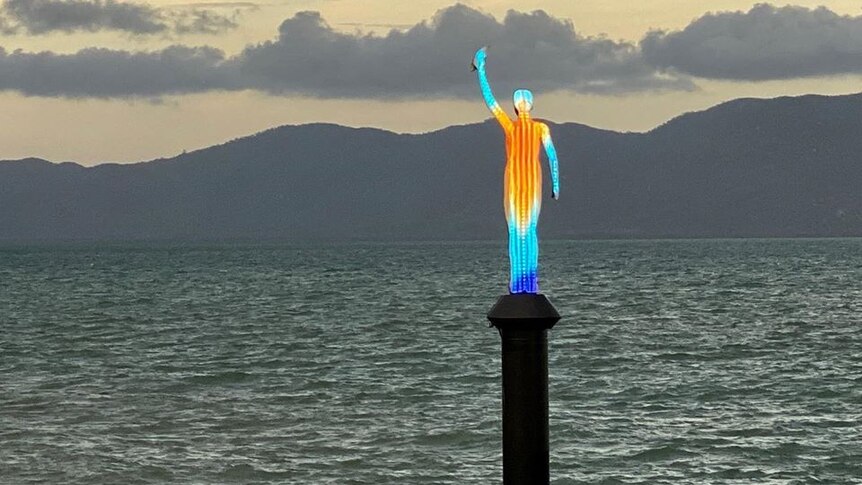 A light sculpture in the shape of a person holding up an arm stands on a post in the middle of deep water.
