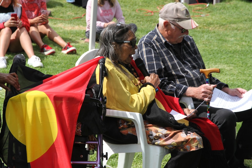 An Aboriginal Elder sits in a chair next to her walker with the Aboriginal flag.