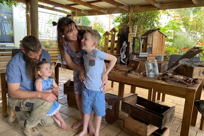 A family in their backyard patio with wooden products on a table and a garden in the background.