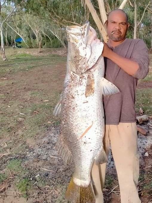 Man wearing brown shirt and beige pants with cigarette in his mouth holds large fish almost as big as him