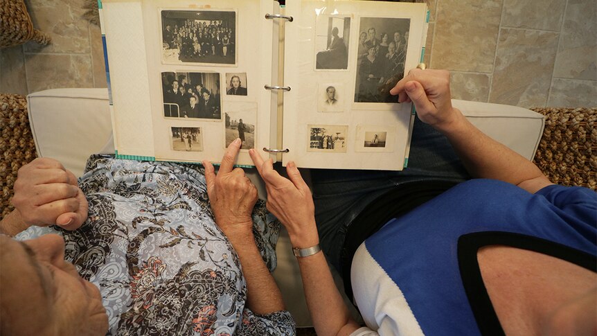 Two women sit on a sofa looking at an album of black and white photos, viewed from above.