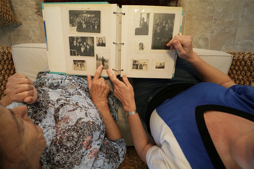 Two women sit on a sofa looking at an album of black and white photos, viewed from above.