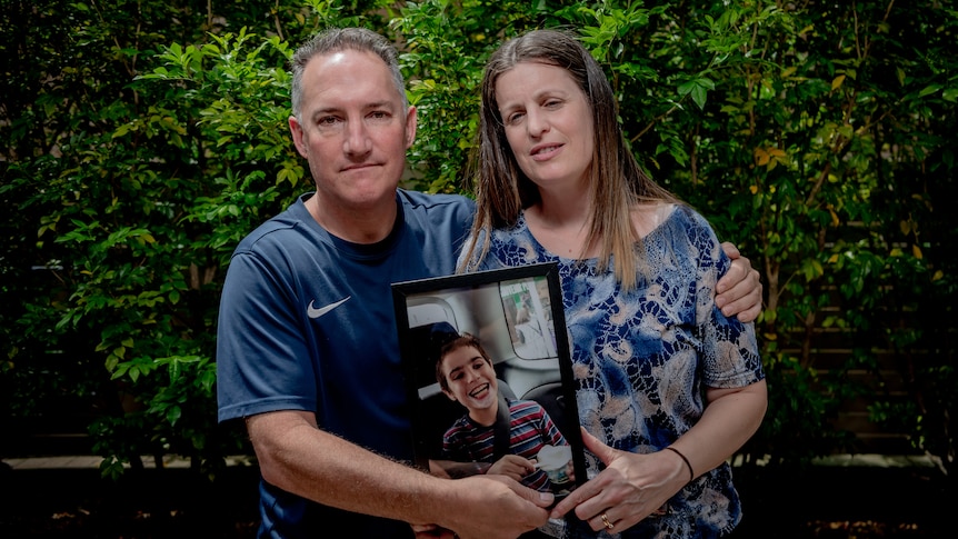 A forlorn man and woman holding a photo of their deceased young child