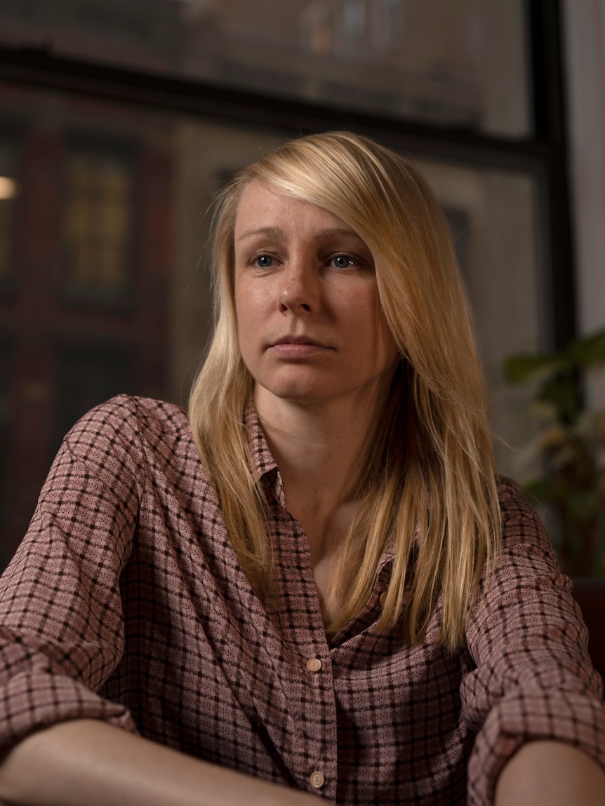 A woman with long blonde hair wears pink check button up shirt looks to the left and sits near apartment window in the evening.