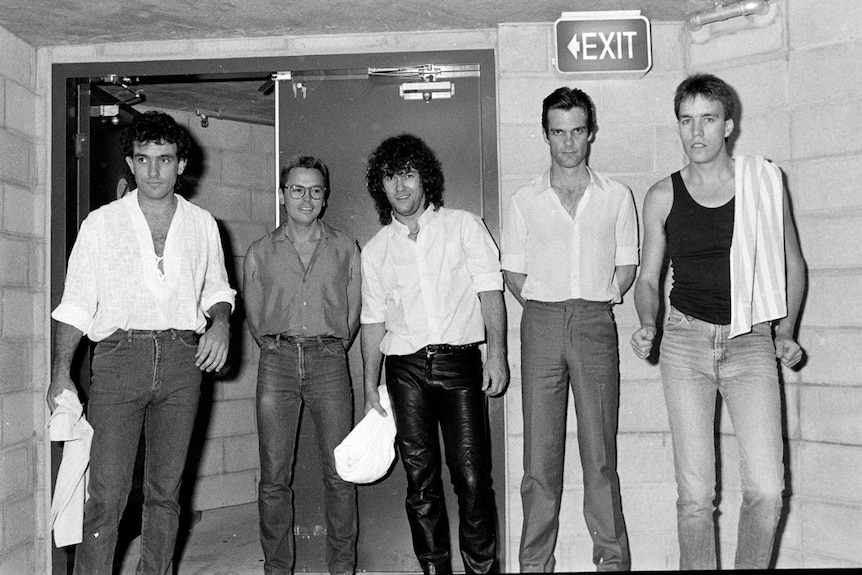 Black and white: Five members of Cold Chisel standing against brick wall backstage 