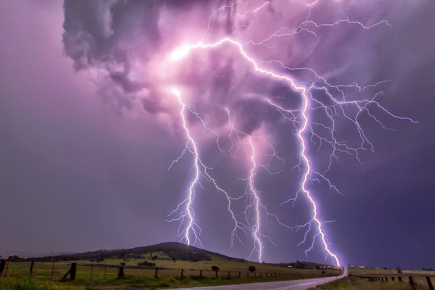 Three lightning bolts strike on two small grassy hills with a road in the foreground.