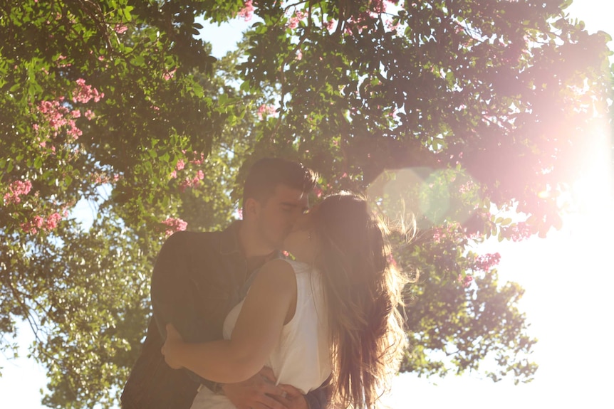 A young couple of high school sweethearts embrace and kiss under a tree dotted with pink flowers.