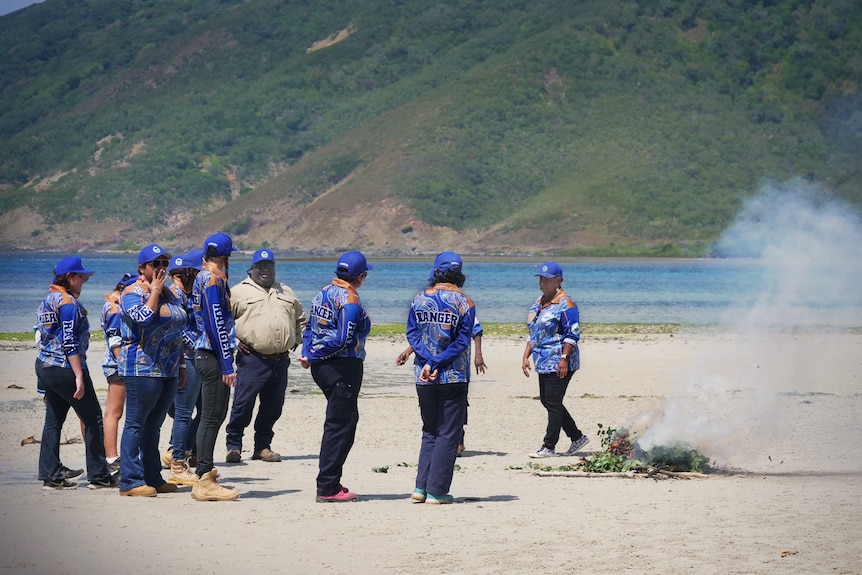 A group of rangers in blue collared shirts and caps stand around a fire on a sandy beach