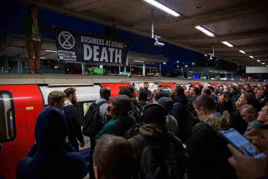 A large crowd stands in front of a train with a man standing on top with banner saying "business as usual = death"