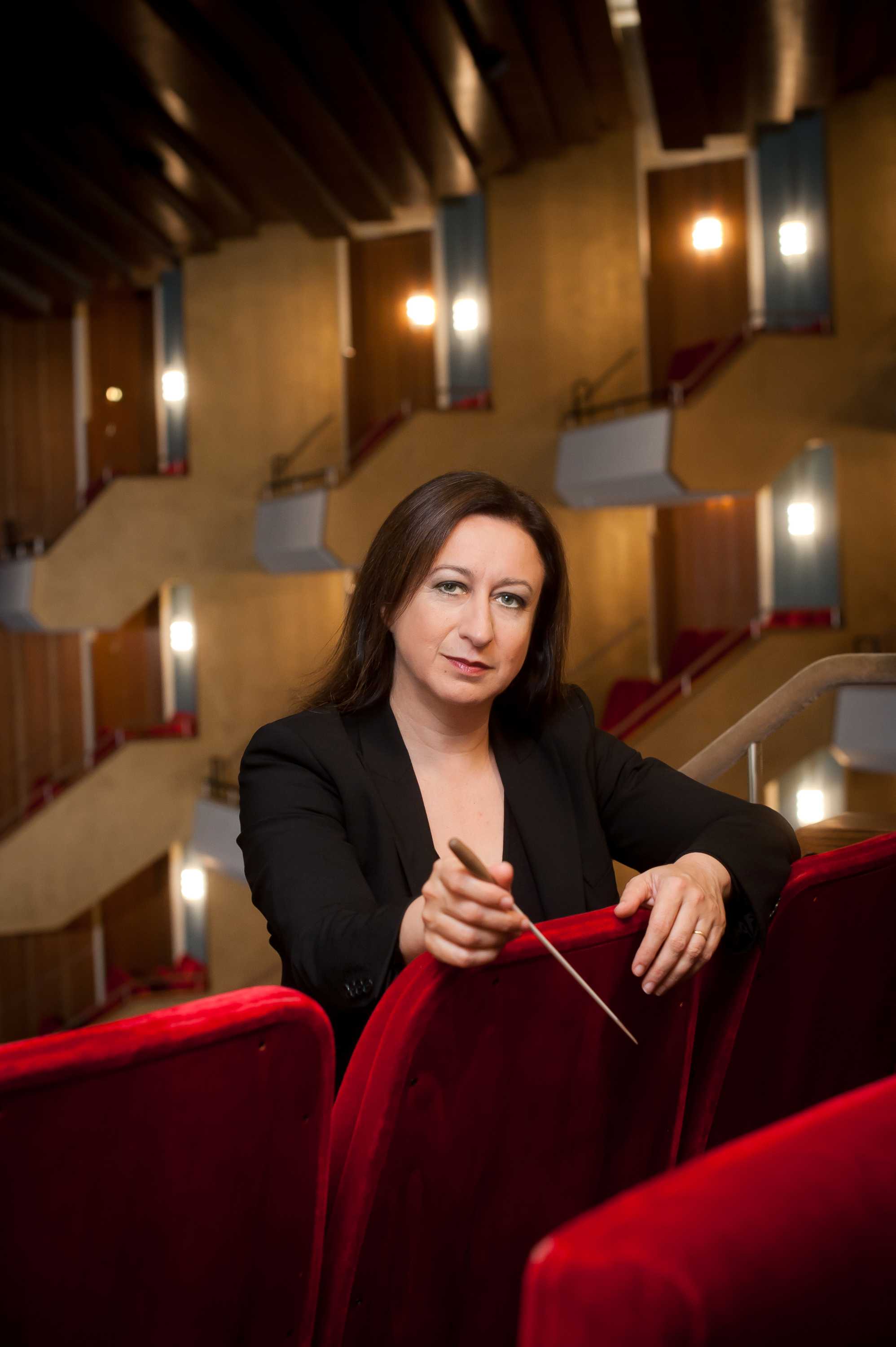 White middle-aged woman with long brown hair wears a black suit and holds a conductor's baton in an auditorium