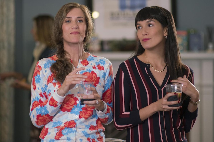A dark haired woman looks to the smug looking fair haired woman beside her in floral cardigan, both hold drinks in plastic cups.