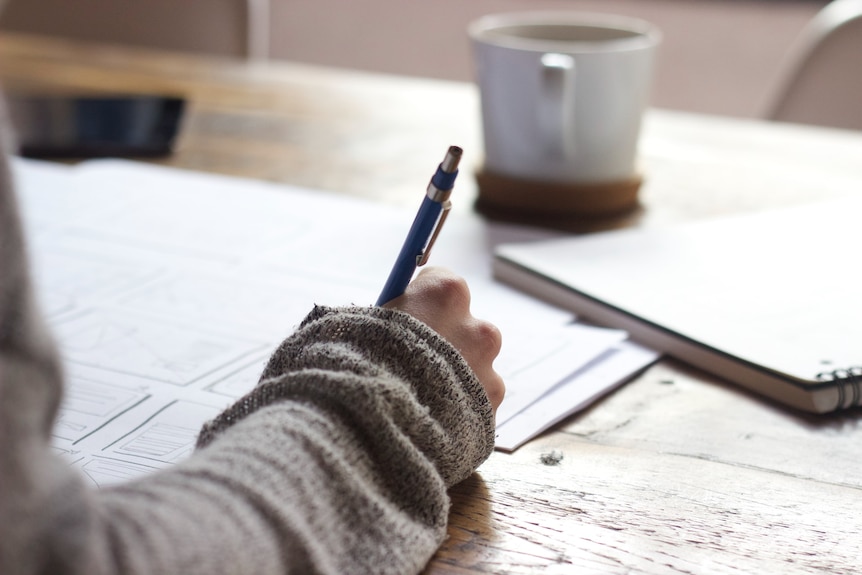 A person with a grey sleeve is writing with a pen on a desk with papers and a white mug.