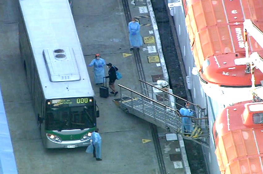 An aerial shot showing a man pushing a suitcase with a facemask walking towards a bus. Blue-gowned and masked people stand near.