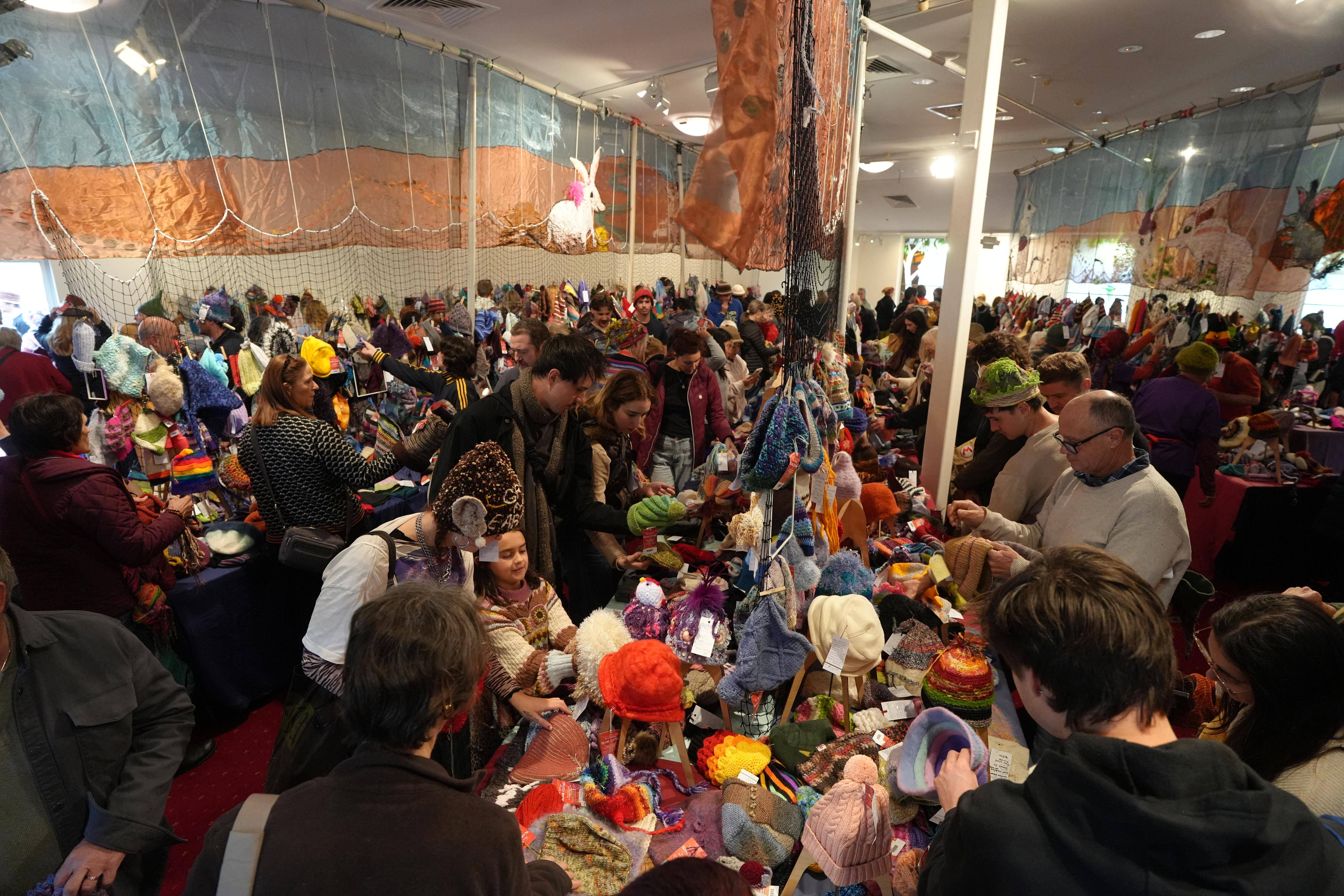A big crowd of people in an indoor large room, most wearing beanies, looking over beanie products laid out on table stalls.