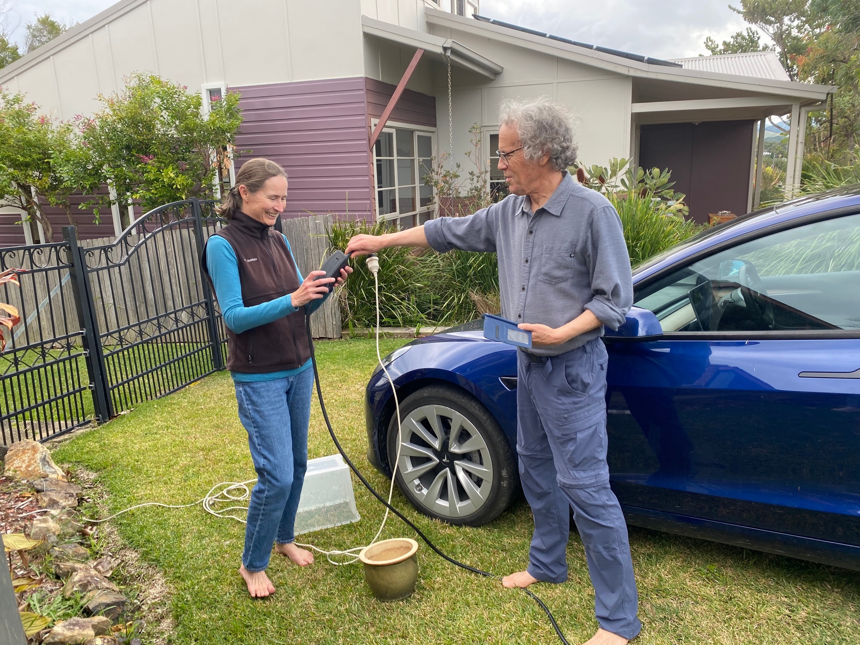 Una pareja parada en su patio delantero configurando un cable de extensión para cargar su Tesla azul. 