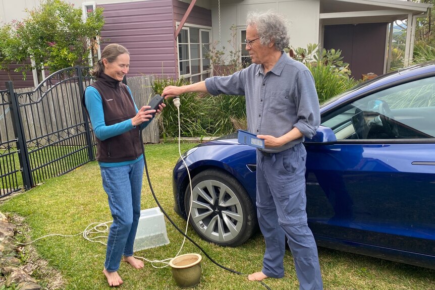 a couple standing in their front yard configuring an extension cord to charge their blue tesla 