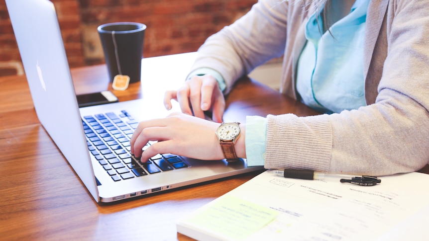 A woman sits at a desk at her laptop, with a book and pen next to her.