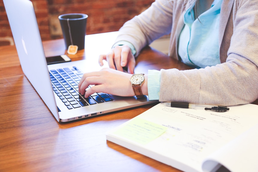 A woman sits at a desk at her laptop, with a book and pen next to her.