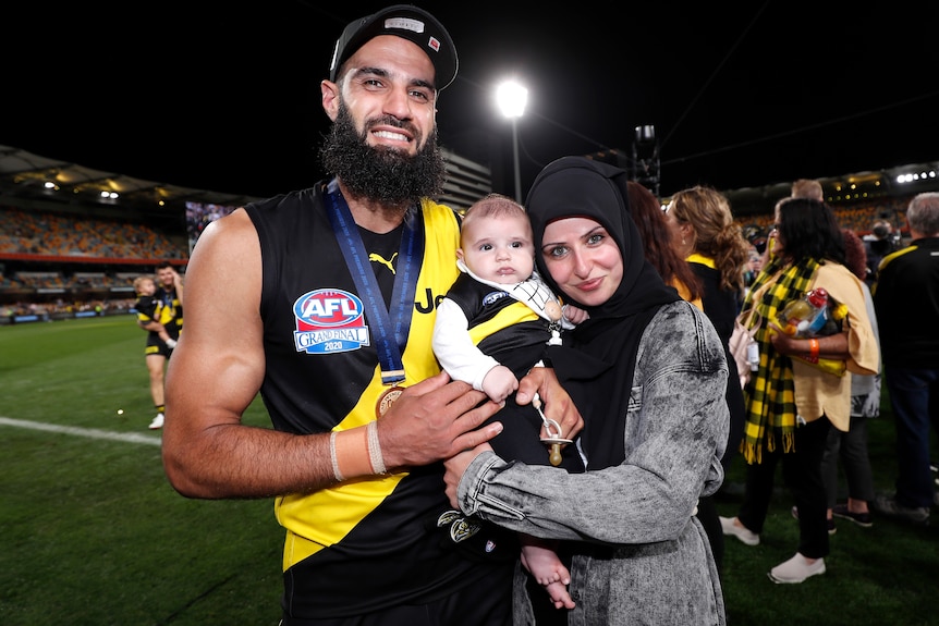 Bachar Houli y su esposa sostienen a su bebé en el Gabba.  Todavía usa su equipo de juego y tiene una medalla alrededor del cuello.