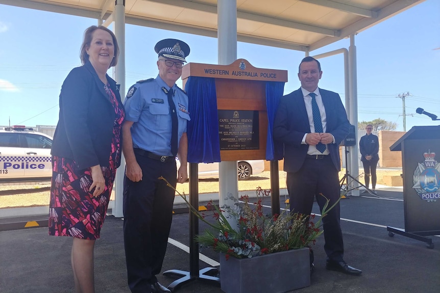 A woman, a police officer, and a man in a suit stand in front of a plaque.