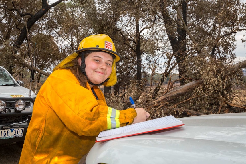 Bec Johns smiles at the camera, pen and notebook in hand leaning on the car bonnet.