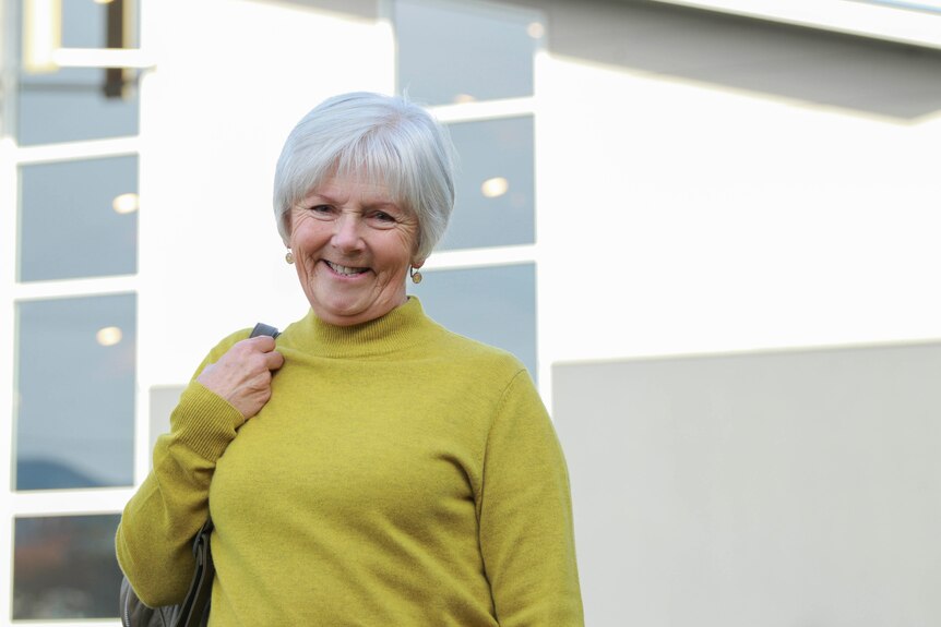A woman with short grey hair stands outside a building, smiling