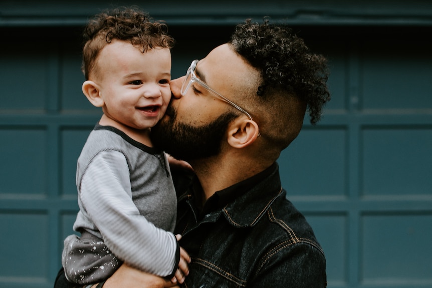 A man with beard and black hair holds his small, laughing child up high and kisses him on the cheek. hi