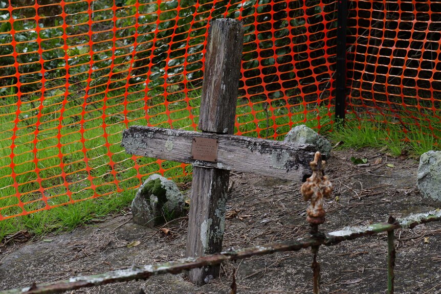 An old wooden cross over a grave site, surrounded by plastic fencing.