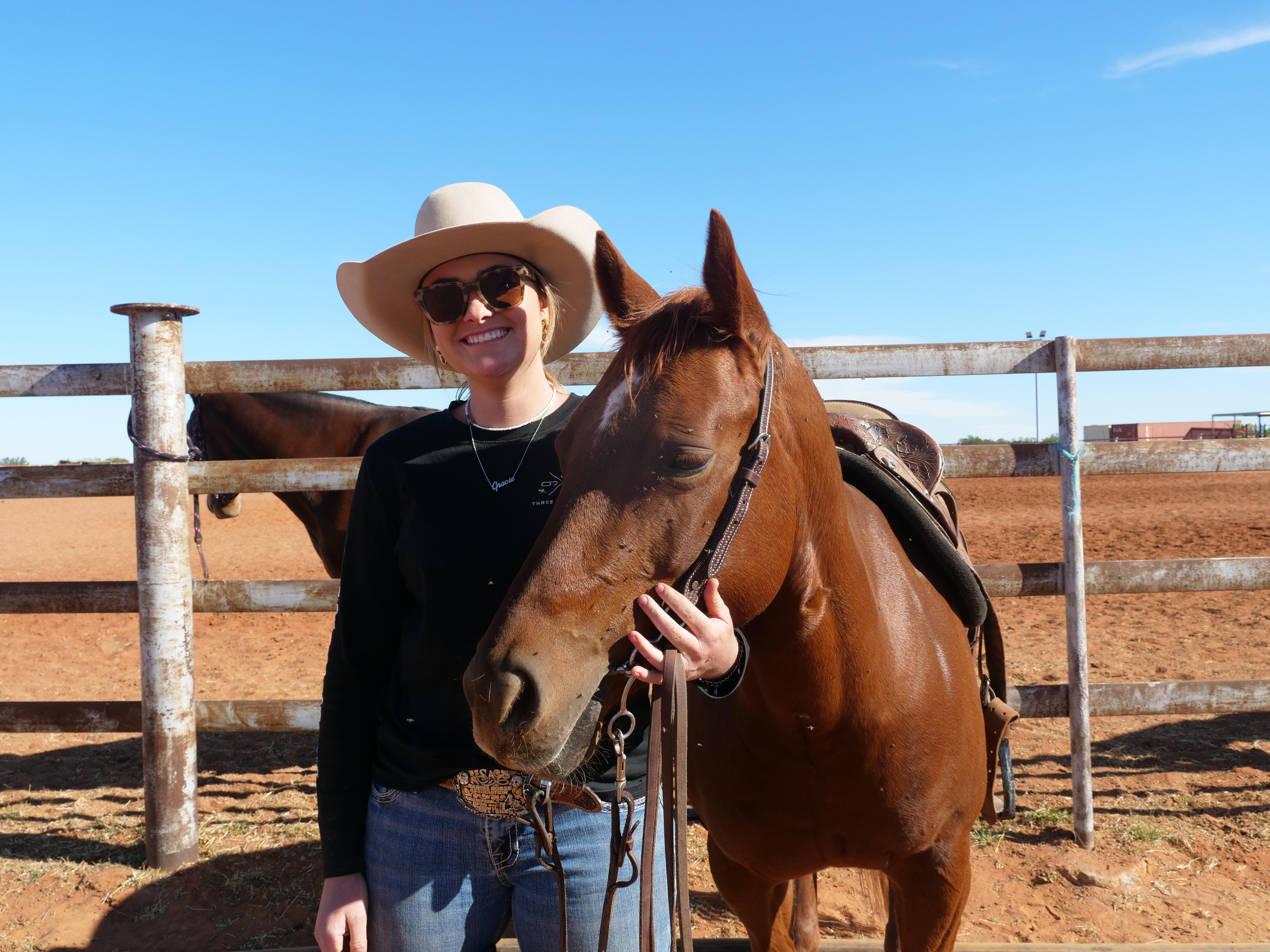 a young girl with sunglasses and a large hat standing next to a brown horse in front of a fence 