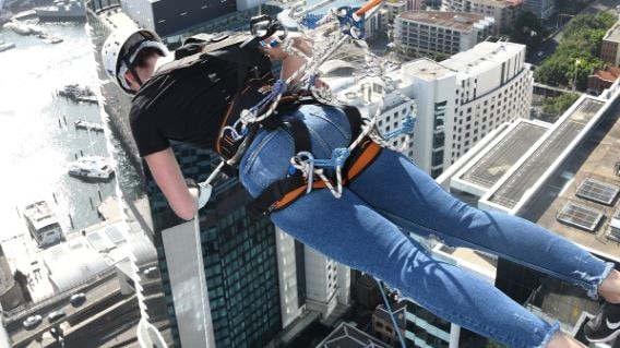 A woman wearing jeans and a helmet is supported by ropes as she leans out from a high-rise building at a 90-degree angle.