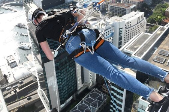 A woman wearing jeans and a helmet is supported by ropes as she leans out from a high-rise building at a 90-degree angle.