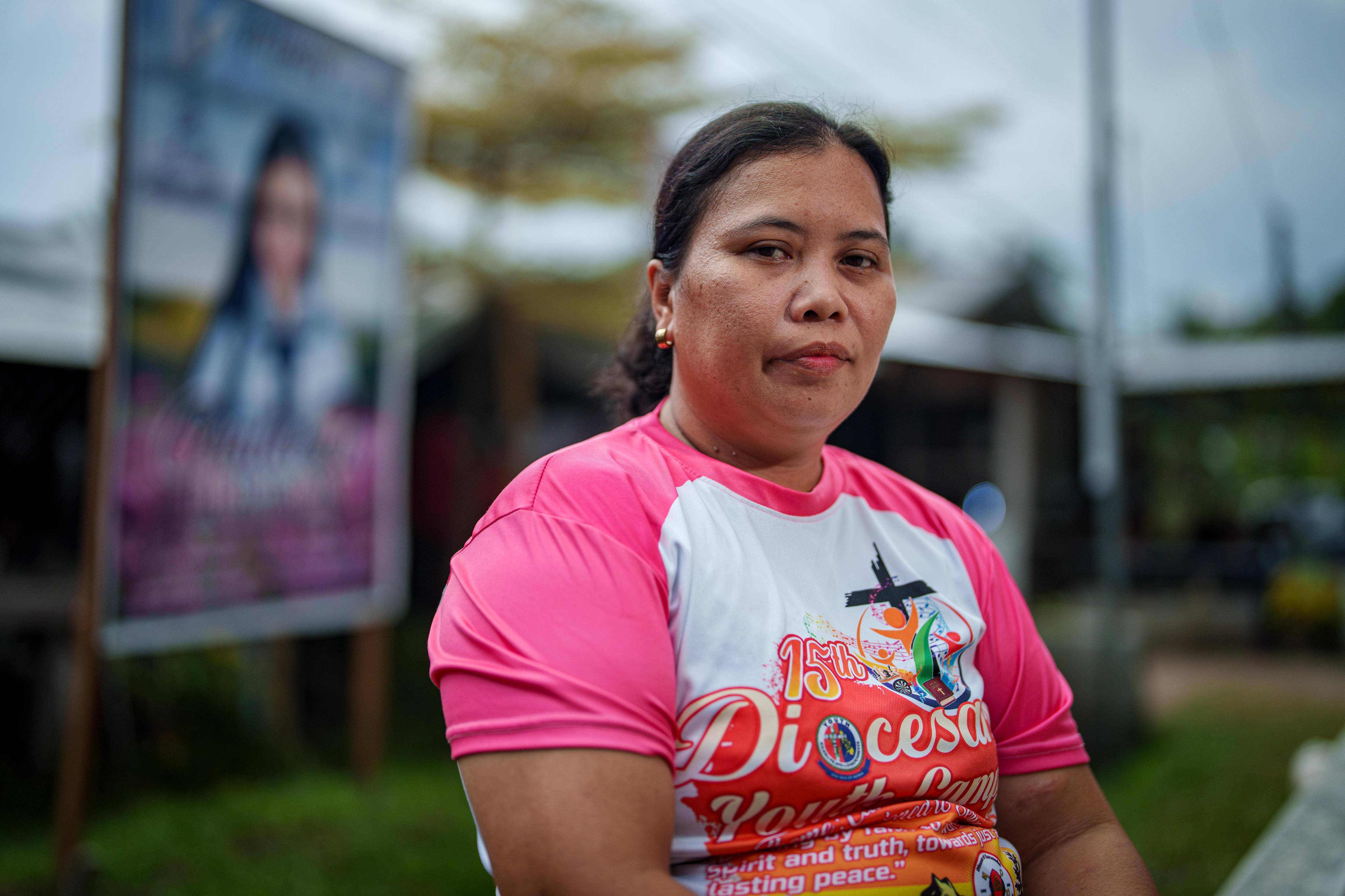 A Filipino woman wearing a pink tshirt outside a small building in a village.
