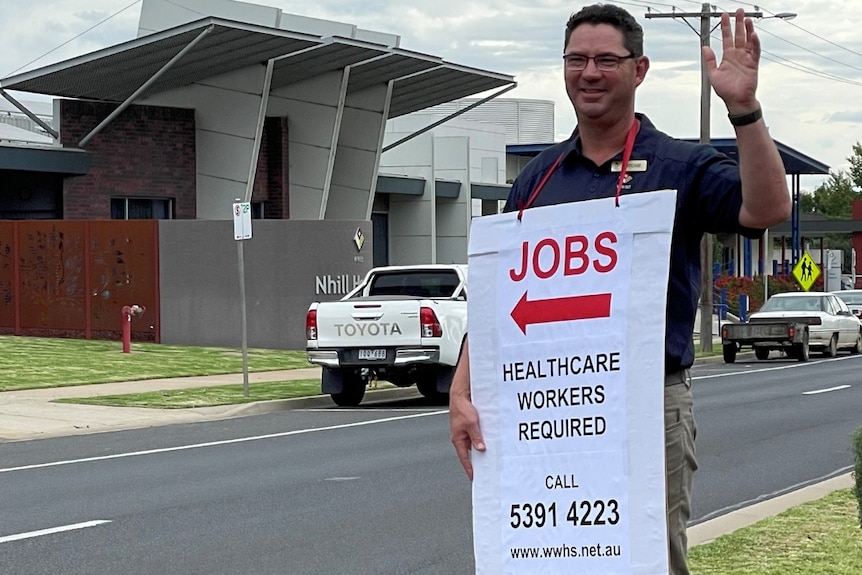A man standing beside the road, waving, wearing a sign saying 'jobs', with an arrow, then 'healthcare workers required'