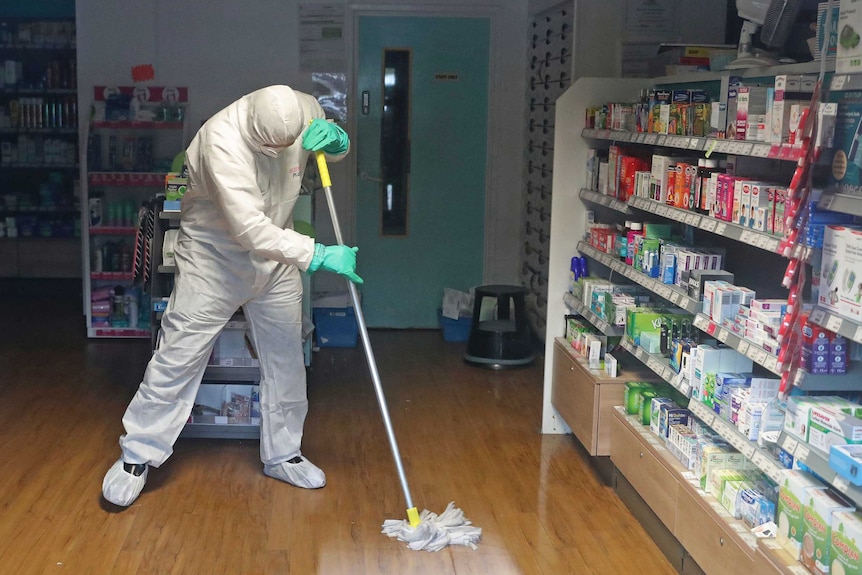 A man in a white hazmat suit wipes a wooden floor with a mop in a pharmacy.