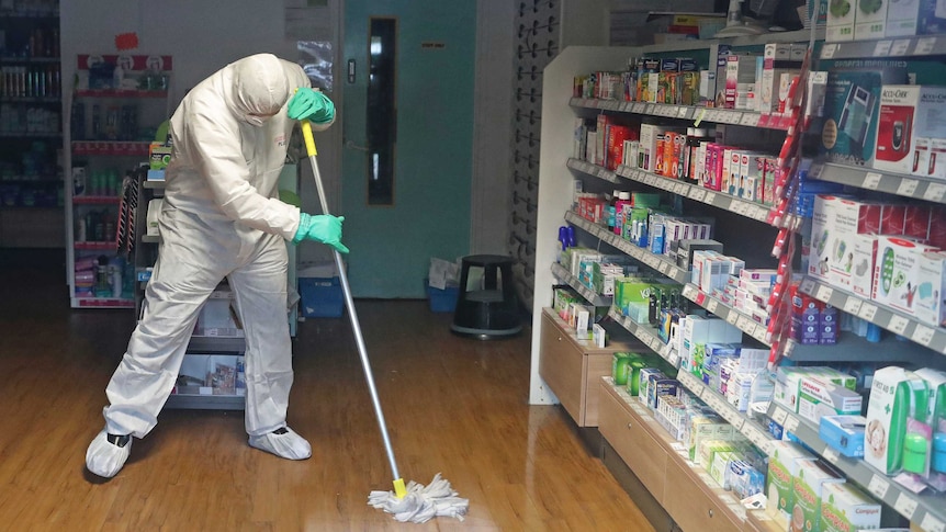 A man in a white hazmat suit wipes a wooden floor with a mop in a pharmacy.