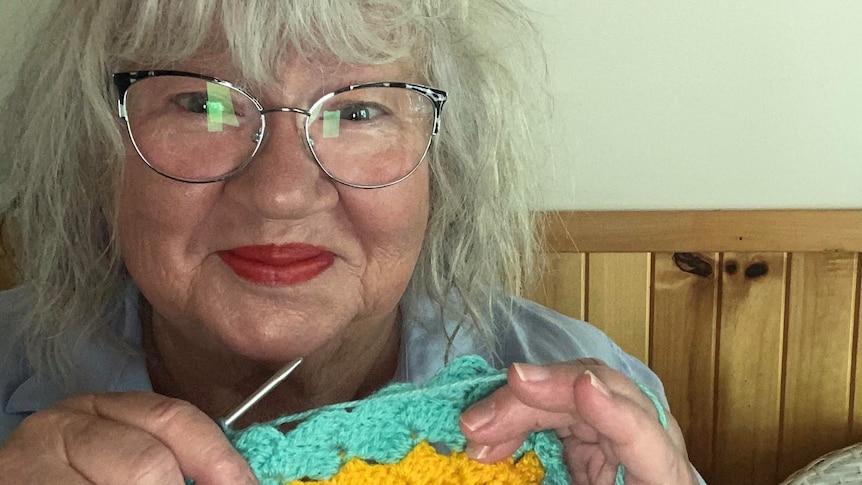 A woman with grey hair, glasses and red lipstick holds a crochet stick and crochet rug in yellow and green