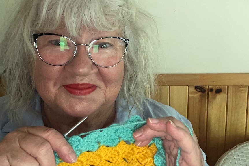 A woman with grey hair, glasses and red lipstick holds a crochet stick and crochet rug in yellow and green