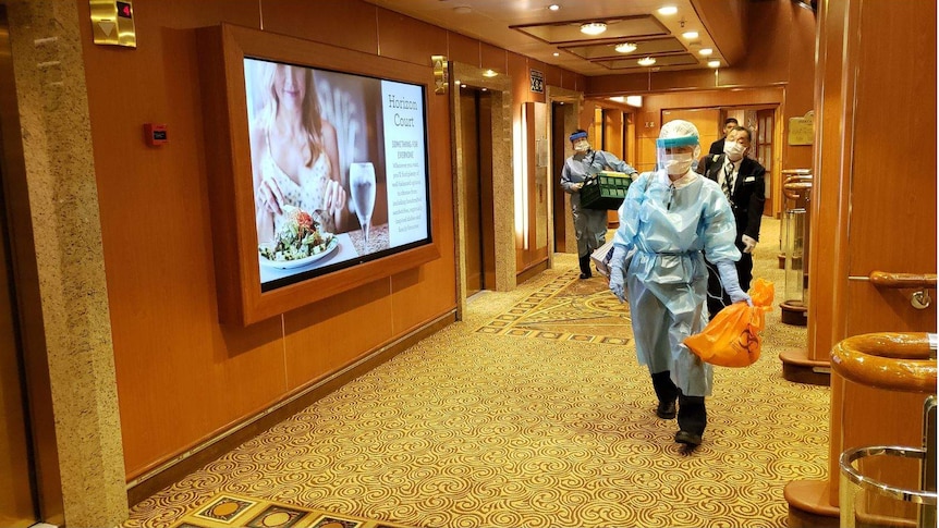 masked health workers clad in blue plastic gowns walking down a corridor on a cruise ship.