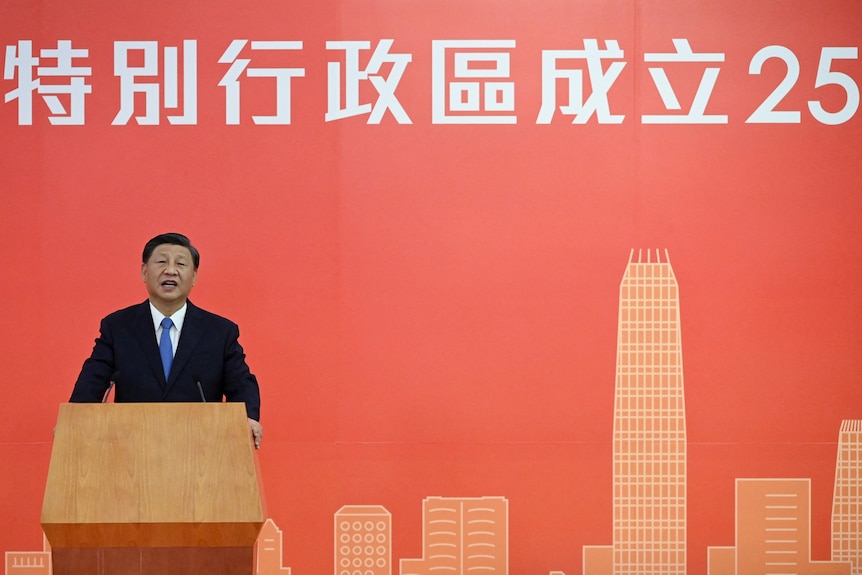 Си Цзиньпин выступает перед кафедрой на красном фоне с написанным на нем числом 25.