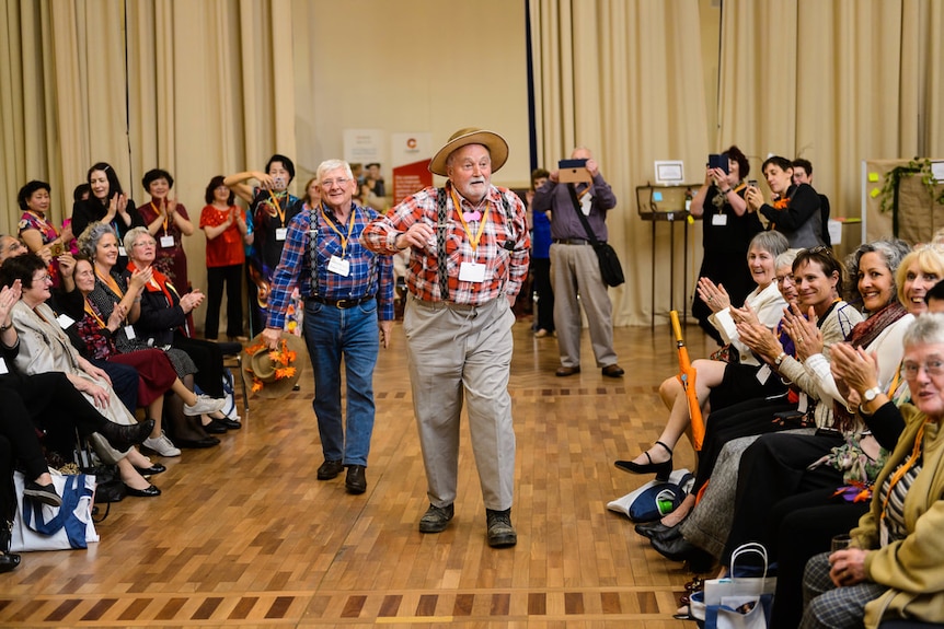 In a hall, two smiling men in flannelette shirts and trousers walk between rows of seated observers, who clap, smile and laugh.