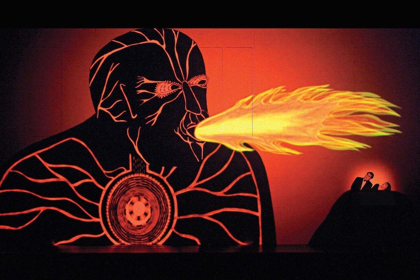 A stage with a projection of a fire breathing man, two people shrink from the fire