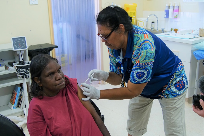 A woman in a blue t-shirt vaccinates another woman in a pink shirt inside a health clinic. 
