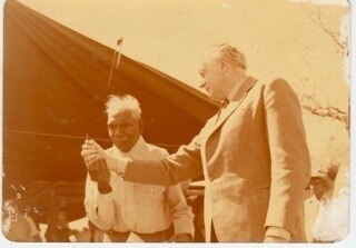 An old photo of a white man pouring sand into the palm of an Aboriginal man.