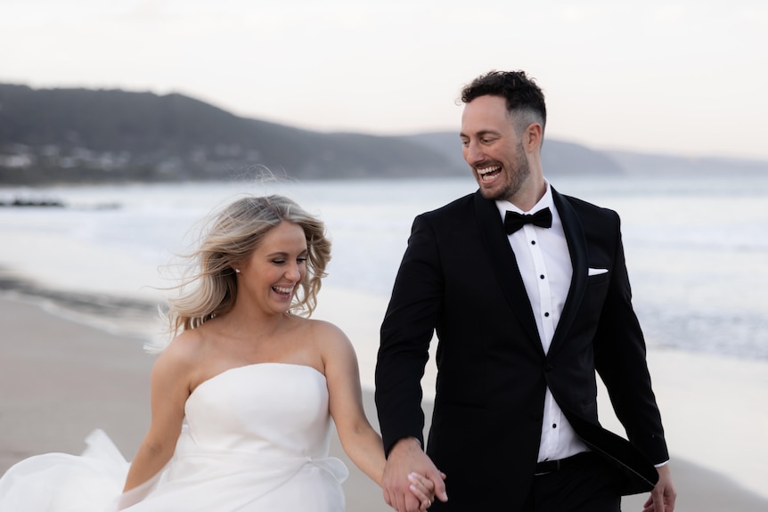 A bride and groom laugh as they walk along a beach, holding hands.