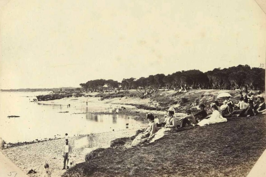 A  black and white photo of a beach. Trees line the hills behind. People in upper-class Victorian-era dress sit in foreground.
