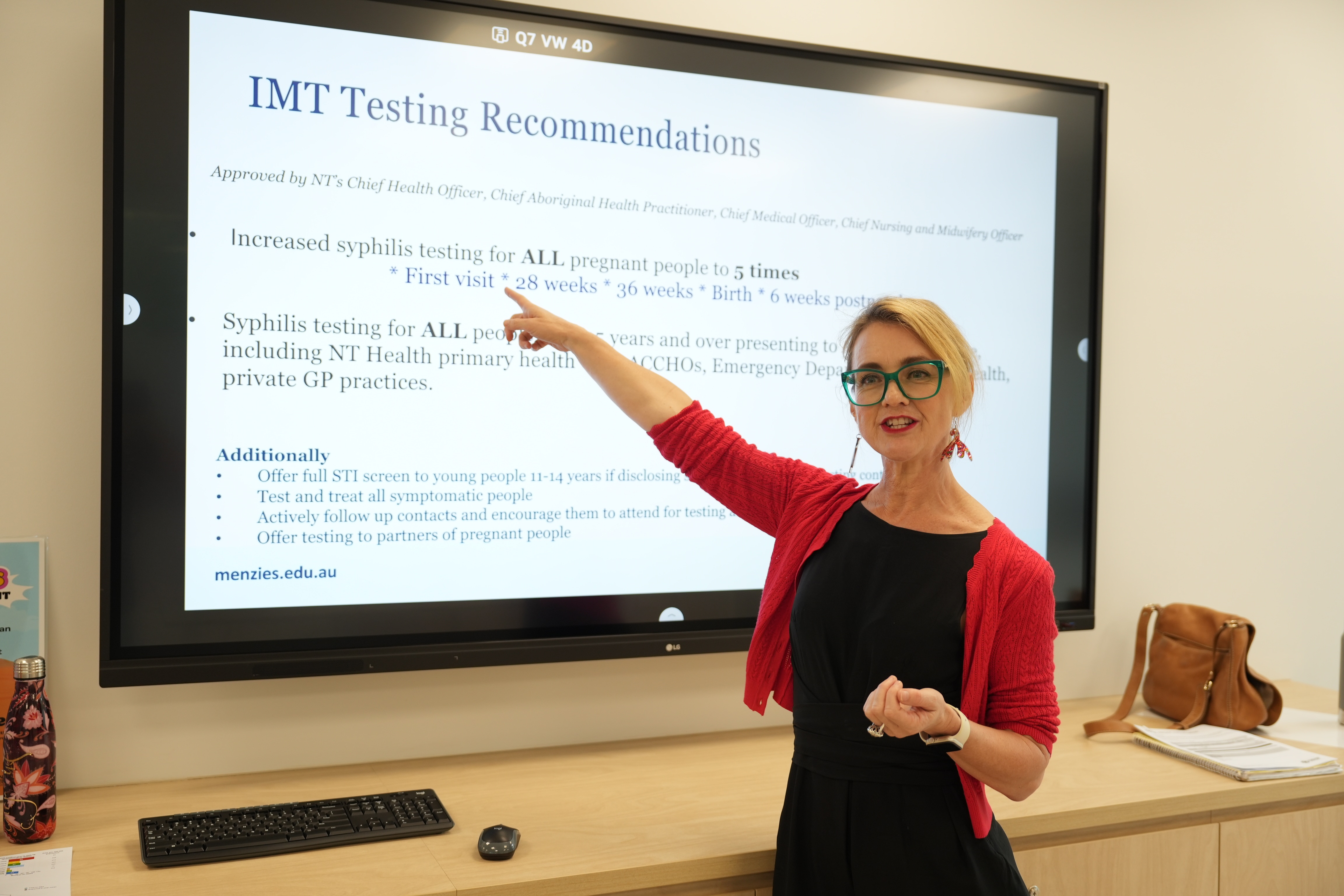 A blonde woman in a red cardigan and glasses points at a screen, which has information about testing for health conditions.