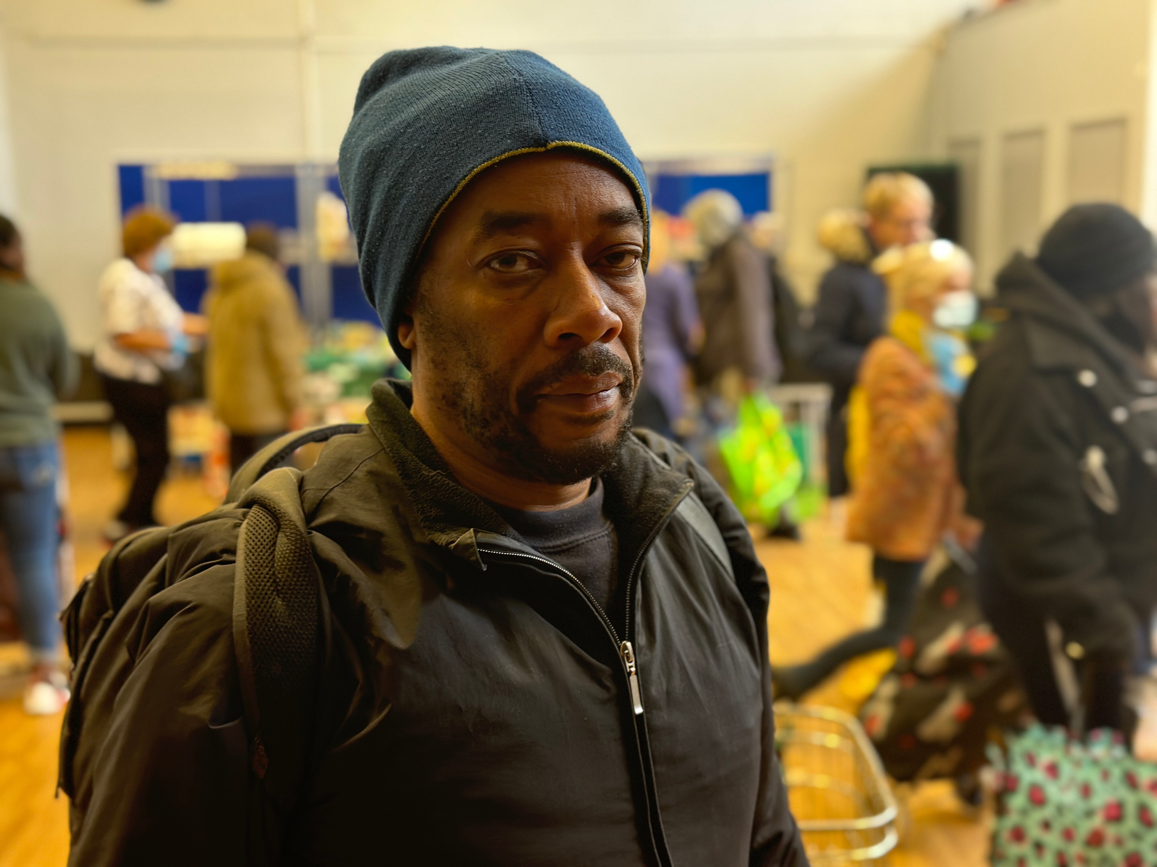 A man in a beanie looks at the camera. Foodbank shoppers mill around him.
