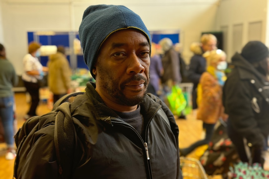 A man in a beanie looks at the camera. Foodbank shoppers mill around him.