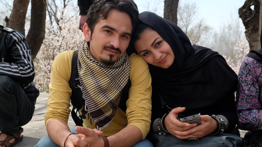 Young dark haired couple sit together. Her head is covered with traditional robes, he wears Western clothes